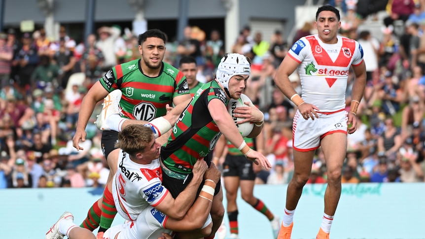 A South Sydney NRL player wearing headgear falls forward carrying the ball as he is tackled by several defenders.