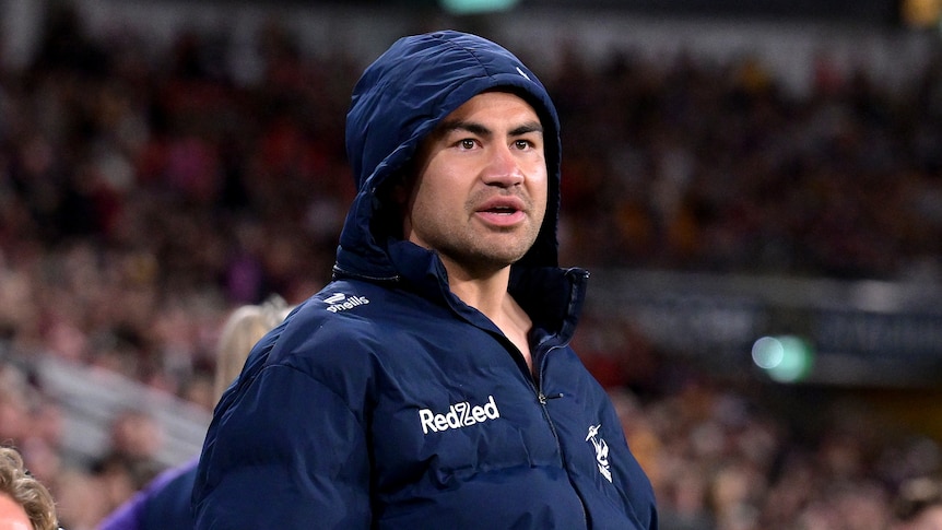 A Melbourne Storm player stands on the sidelines wearing a jacket with a hood on.