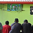 Seven men stand at a table watching a rugby league match on TV.