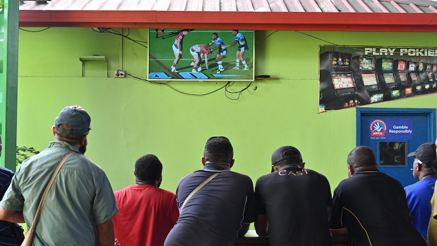 Seven men stand at a table watching a rugby league match on TV.